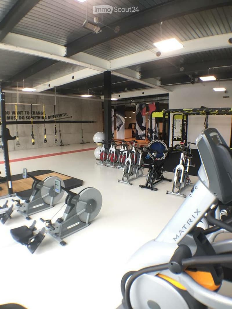 Modern fitness studio with various exercise equipment including stationary bikes, elliptical machines, treadmills, and weight machines. The space features a high ceiling with exposed beams and ample lighting.