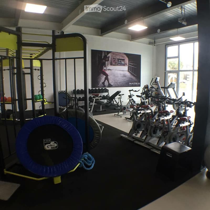 Well-lit gym room with various exercise equipment including treadmills, elliptical machines, weight racks, and free weights. Features a large poster on the wall and a foam roller on the floor.
