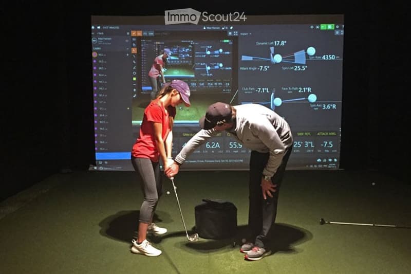 Two individuals practice golf on an indoor simulator, using a golf club and observing data on a large screen.