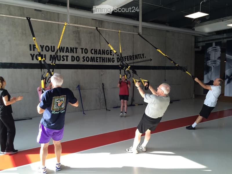 A group of people are exercising in a gym using TRX suspension trainers. The gym has a motivational quote on the wall and a clean, spacious layout.