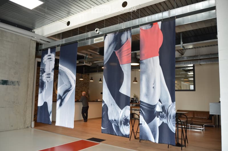 Modern, open-plan space with exposed metal beams and large banners featuring fitness imagery. Includes a sofa, several stools, and a small table.