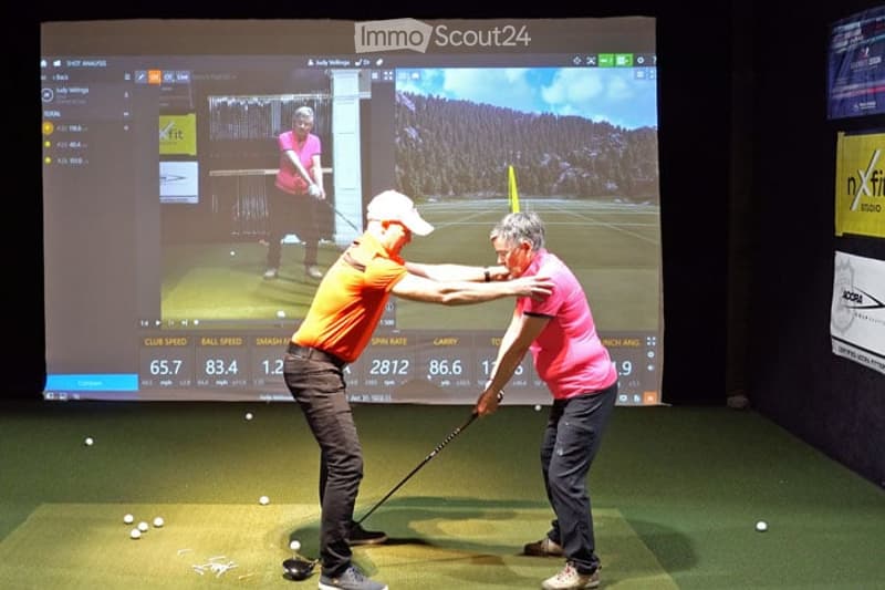 Indoor golf simulator with a large screen displaying a virtual course. Two individuals are practicing golf swings, with one guiding the other. The room has artificial turf and several golf balls scattered on the floor.