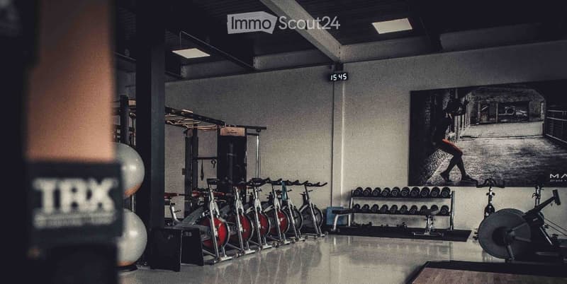 Modern gym space with multiple exercise bikes, dumbbell rack, fitness ball, and weight machine. Large black-and-white poster on wall.