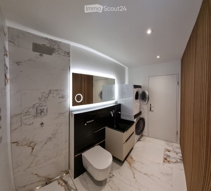 Modern bathroom with marble tiles, a toilet, sink, mirror, shower, and stacked washer/dryer unit.