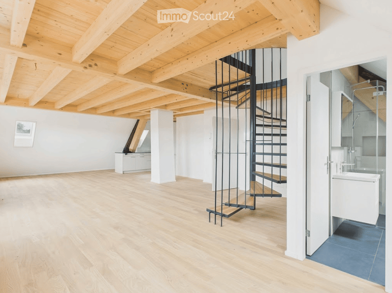 Open-plan space with exposed wooden beams, light wooden flooring, a modern spiral staircase, and a small bathroom area with a bathtub and glass shower enclosure.