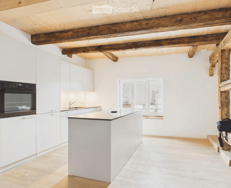 Modern kitchen with white cabinets, a central island, and exposed wooden beams on the ceiling. Features include an oven, sink, and ample natural light from a large window.