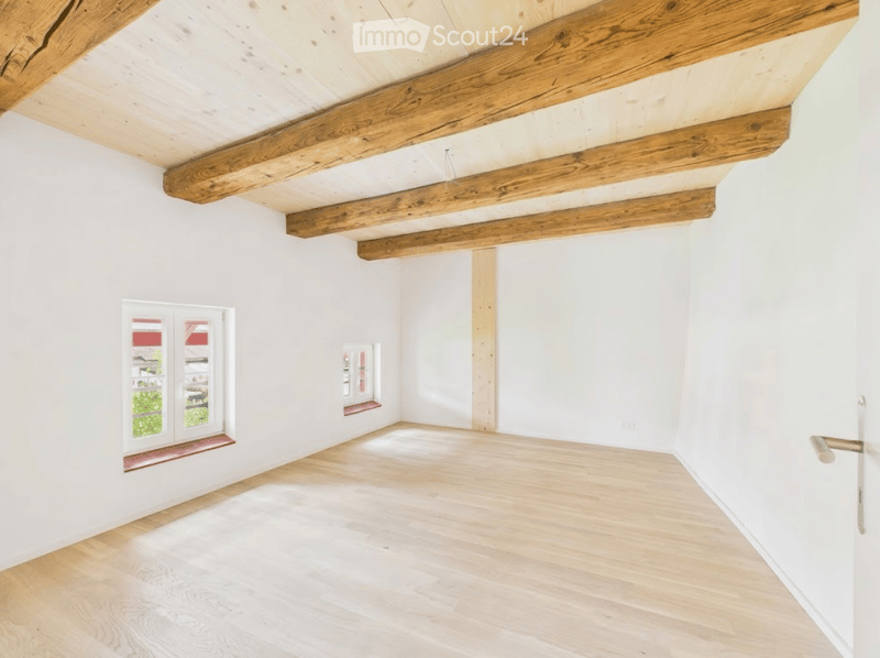 Empty room with white walls, wooden floor, and exposed wooden beams on the ceiling. Two windows with red window sills are visible.