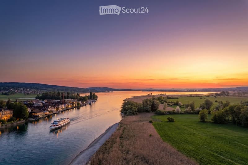 A scenic view of a river at sunset, with a boat on the water, a small town on one bank, and green fields on the other.