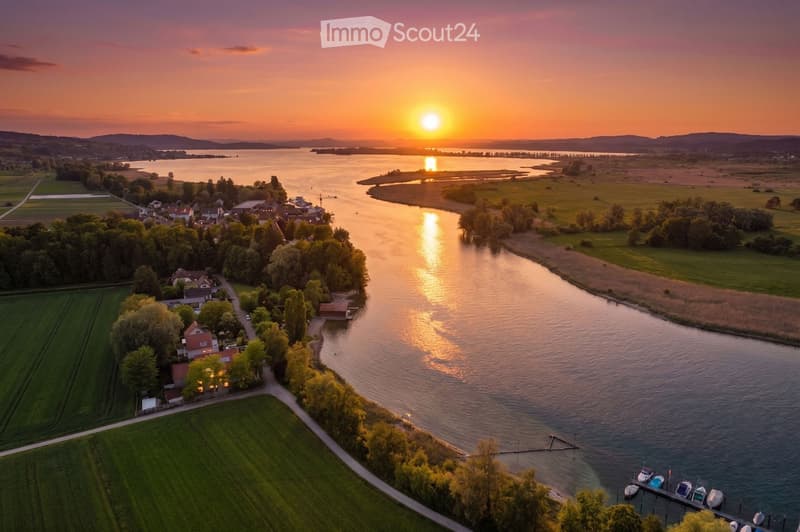 Aerial view of a serene lakeside village at sunset, featuring houses, a dock with boats, and surrounding greenery.