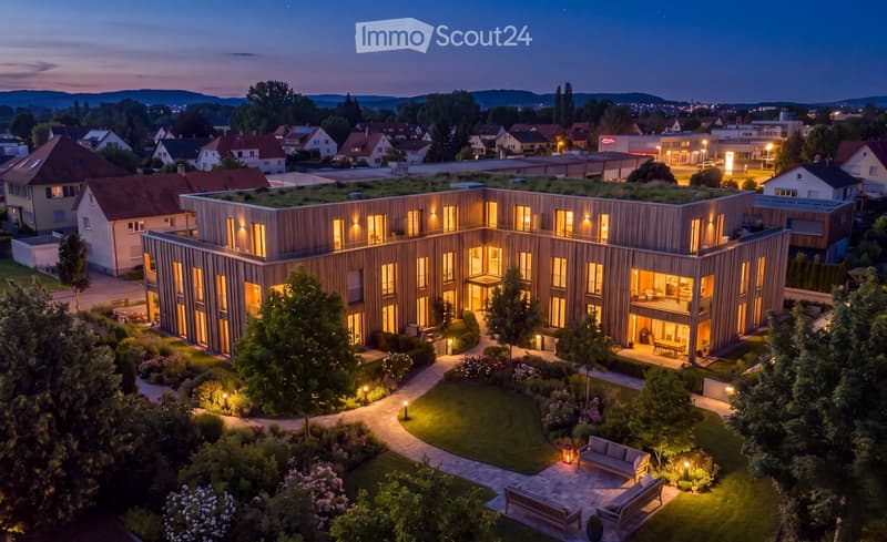 Modern multi-story building with wooden facade and large windows, surrounded by a landscaped garden with pathways, seating areas, and outdoor furniture.