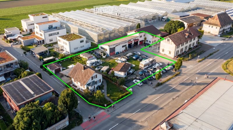 Aerial view of a residential and commercial area with multiple buildings, parking lots, greenhouses, and surrounding greenery.