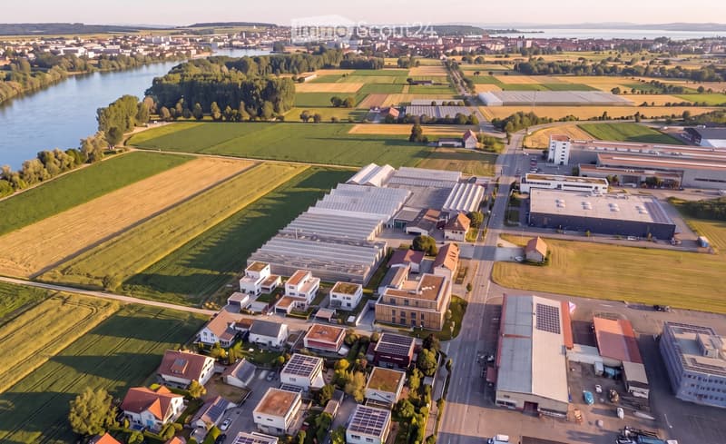 Aerial view of a mixed-use area with residential houses, industrial buildings, greenhouses, and agricultural fields, surrounded by a river and rural landscape.