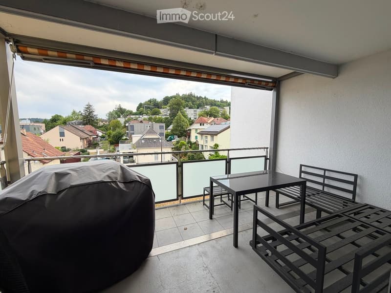 Balcony with a covered grill, a small table with chairs, and a bench, overlooking a residential area with houses and trees.