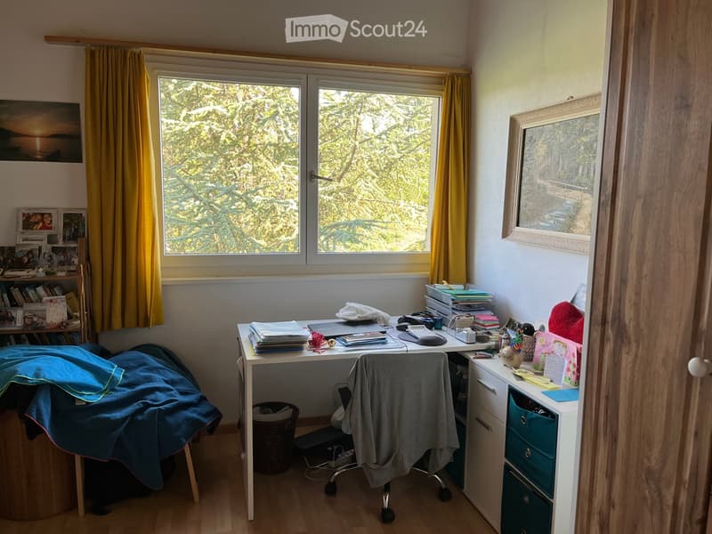 A well-lit room with a desk, chair, laptop, books, framed picture, and curtain. The room has a large window with a view of trees.