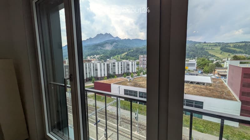 Balcony with metal railing, overlooking a cityscape with buildings, train tracks, and distant mountains.