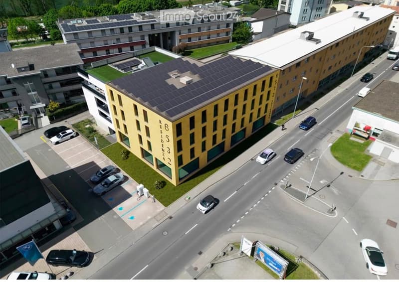 A modern multi-story building with solar panels on the roof, surrounded by a parking area with several cars and a landscaped green space.