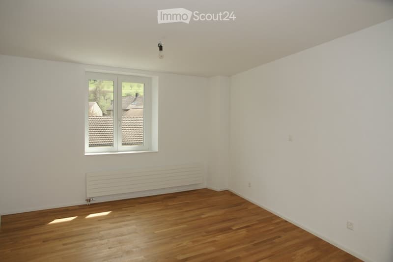 Empty room with wooden flooring, white walls, a large window with white frame, and a radiator below the window.