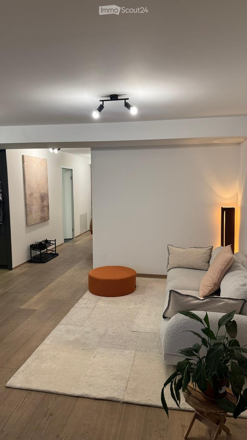 Modern living room with a white sofa, orange stool, small plant on a wooden table, and artwork on the wall.