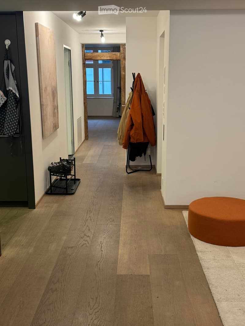A modern hallway with light wooden flooring, white walls, and recessed lighting. Features include a coat rack with jackets, a shoe rack, and a small round orange stool near the entrance.
