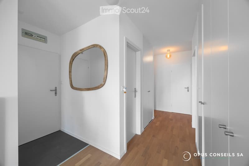 Clean, modern hallway with white walls, wooden flooring, and several doors. A decorative mirror with a wooden frame is mounted on one wall, and a light fixture is visible on the ceiling.