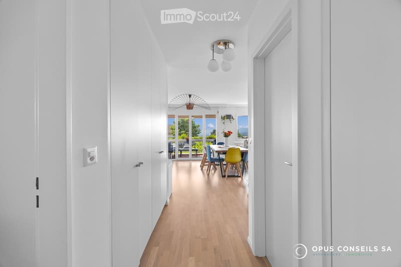Bright, modern hallway with white walls, wooden flooring, and recessed lighting. Opens into a dining area with a table, chairs, and a view of a garden.