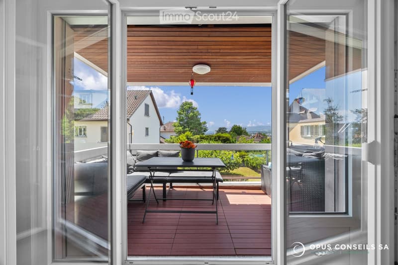 Balcony with wooden decking, glass sliding doors, outdoor furniture including a table, chairs, sofa, and a flower pot, overlooking a scenic view of houses and greenery.