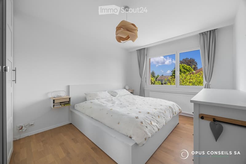 Bright bedroom with white walls, wooden flooring, large windows with grey curtains, a white bed with patterned bedding, a nightstand with a lamp, and a hanging pendant light.
