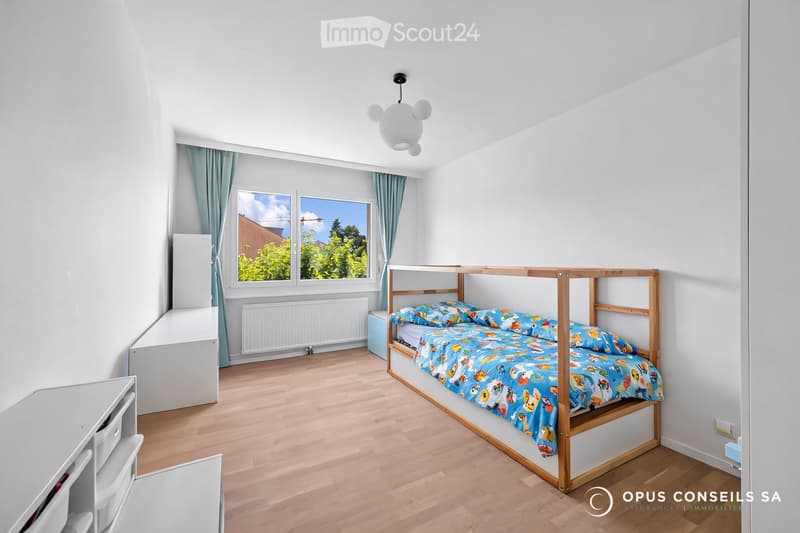 Bright bedroom with a wooden bunk bed, colorful bedding, white walls, wooden flooring, and large windows with blue curtains.