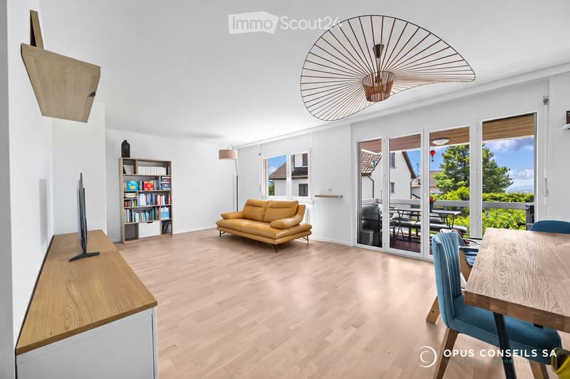 Bright living room with light wooden flooring, a yellow leather sofa, a wooden dining table with blue chairs, a bookshelf, and a television on a wooden stand. Large glass doors lead to a balcony with outdoor furniture visible.