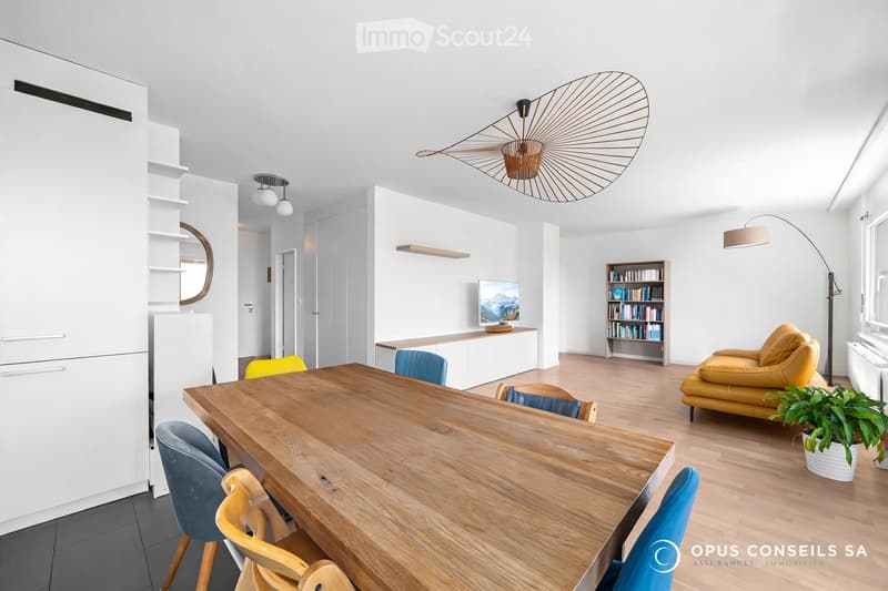 Bright, modern living and dining area with white walls, wooden flooring, and contemporary furniture including a sofa, dining table, chairs, bookshelf, and a screen. Features include a decorative wall-mounted light fixture and a floor lamp.