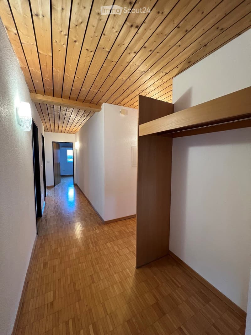A narrow hallway with wooden flooring and white walls, featuring built-in wooden shelving on one side and a light fixture on the wall.