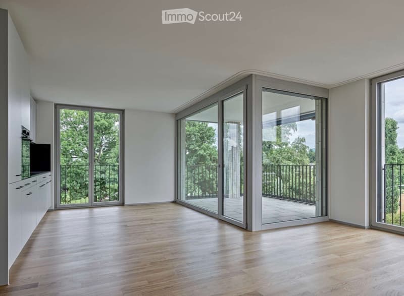 Bright, open-plan room with light wooden flooring, white walls, and large sliding glass doors leading to a balcony with a view of greenery.