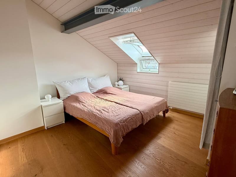 Cozy attic bedroom with wooden floors, slanted ceilings, and a skylight window. Features a double bed with white pillows and a pink duvet, a white nightstand, and a radiator.
