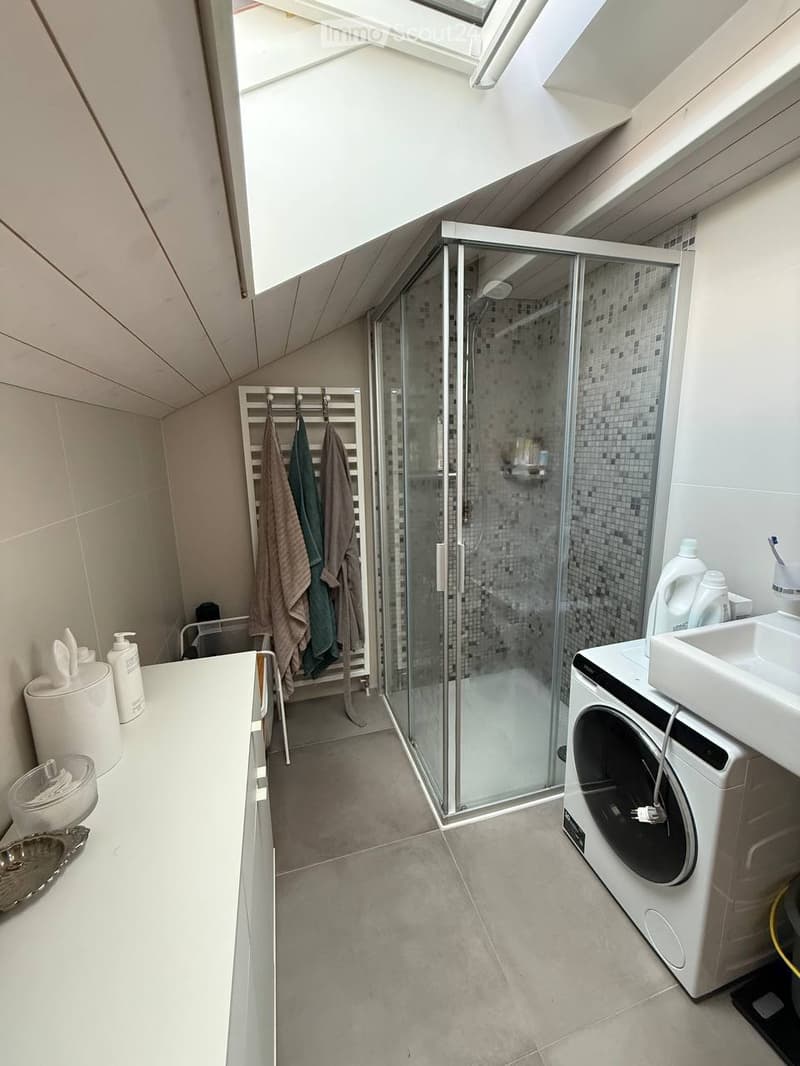 Modern bathroom with a glass-enclosed shower, white sink, washing machine, and towel rack with towels. The room features a skylight and neutral-colored tiles.