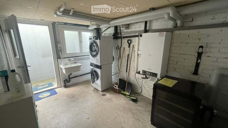Utility room with a washing machine, sink, broom, mop, and a small fridge. Exposed brick wall and concrete floor.