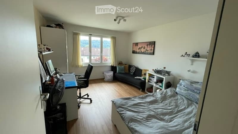 Cozy bedroom with a double bed, a small sofa, a desk with a chair, a fan, and a view through a window. The room features wooden flooring and white walls.