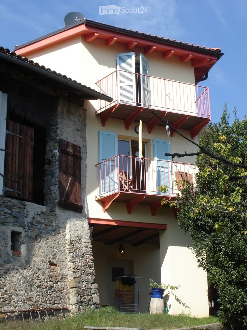 Two-story house with light yellow walls, red trim, and multiple balconies with metal railings. Features include a stone wall section, wooden shutters, and a small garden area with a potted plant.