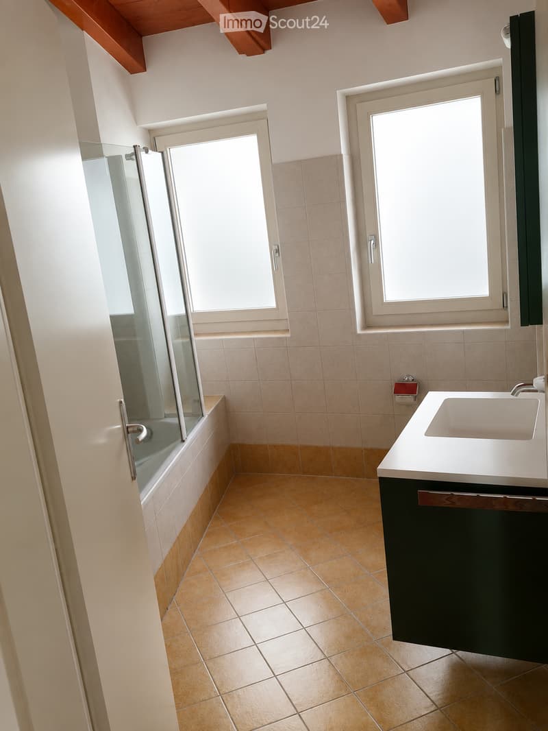 Modern bathroom with beige tiles, a white sink with a black cabinet, a bathtub with a glass shower enclosure, and two windows providing natural light.