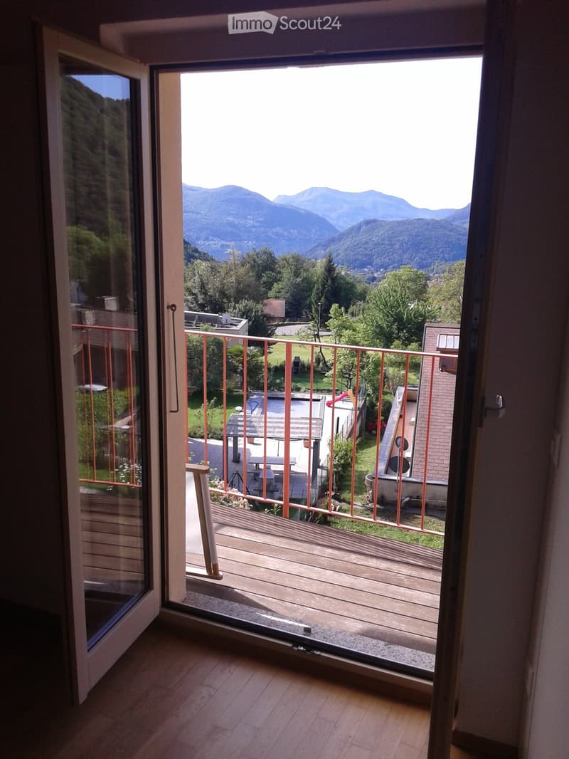 Open sliding glass door leads to a wooden balcony with red metal railing, overlooking a scenic mountain view and a landscaped garden area with a table and chairs.