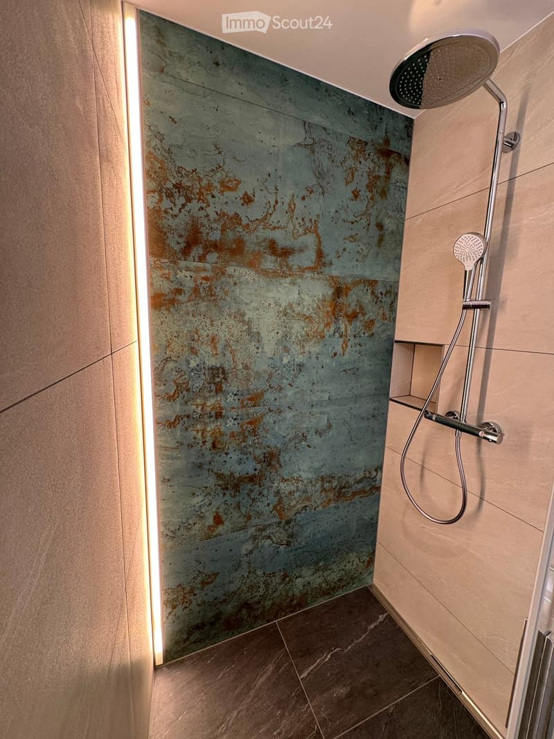 Modern bathroom shower area with a sleek, metallic showerhead and hose, mounted on a wall with a textured, two-tone tile design in green and brown.
