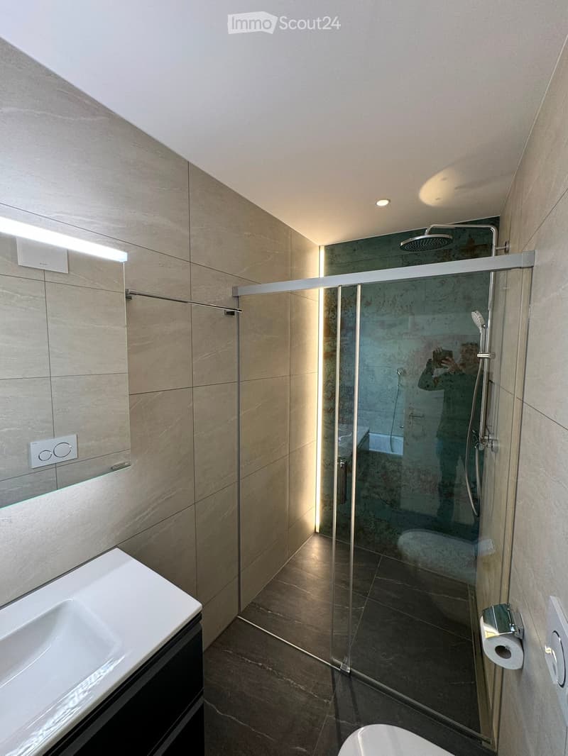 Modern bathroom with a glass-enclosed shower, a white sink, and a toilet. The walls are tiled in beige marble, and the floor is dark tiled.