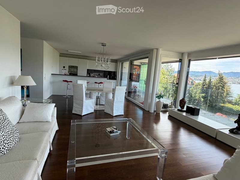 Modern living room with a sofa, coffee table, dining area with chairs and stools, large windows offering a scenic view, and decorative items including a vase and statue.
