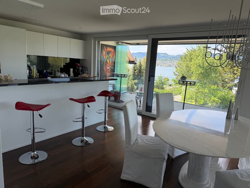 Modern kitchen and dining area with white cabinets, a black countertop, red bar stools, a white dining table with chairs, and a view of a lake and mountains through large windows.