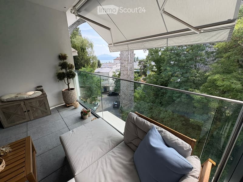 Balcony with a modern sofa, cushions, a potted plant, and a wooden cabinet, offering a scenic view of the city and mountains.