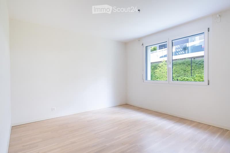 Empty room with white walls and light wooden flooring, featuring a large window with a view of greenery outside.