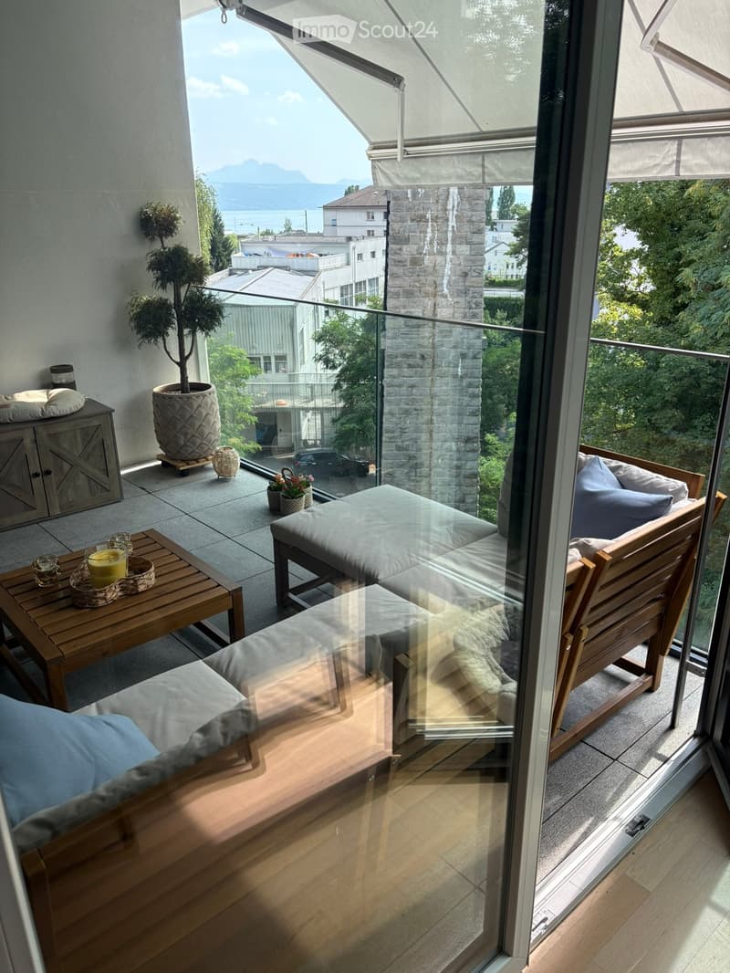 Balcony with a view of the lake and mountains, featuring a sofa, a small table, a chair, and a potted plant.