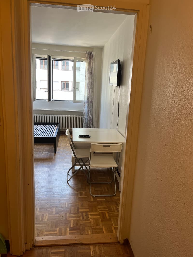 Small furnished room with wooden flooring, a bed, a table with two chairs, and a television mounted on the wall. The room has large windows with open shutters, allowing natural light to enter.