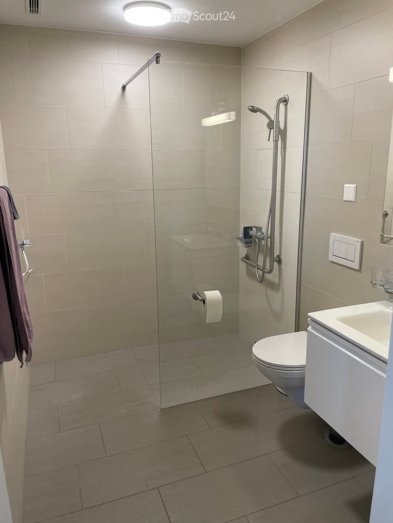 Modern bathroom with beige tiles, a glass-enclosed shower, a toilet, a sink, and a towel hanging on the wall.