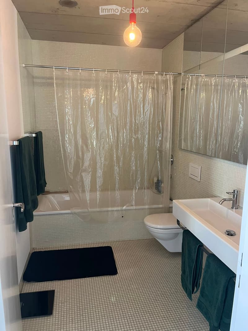 Modern bathroom with a bathtub, toilet, sink, mirror, and green towels. Features a shower curtain and a dark bath mat on the floor.