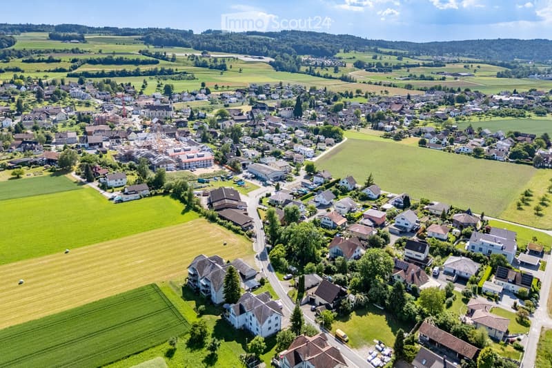 Aerial view of a small village with numerous houses, green fields, and a church in the background.